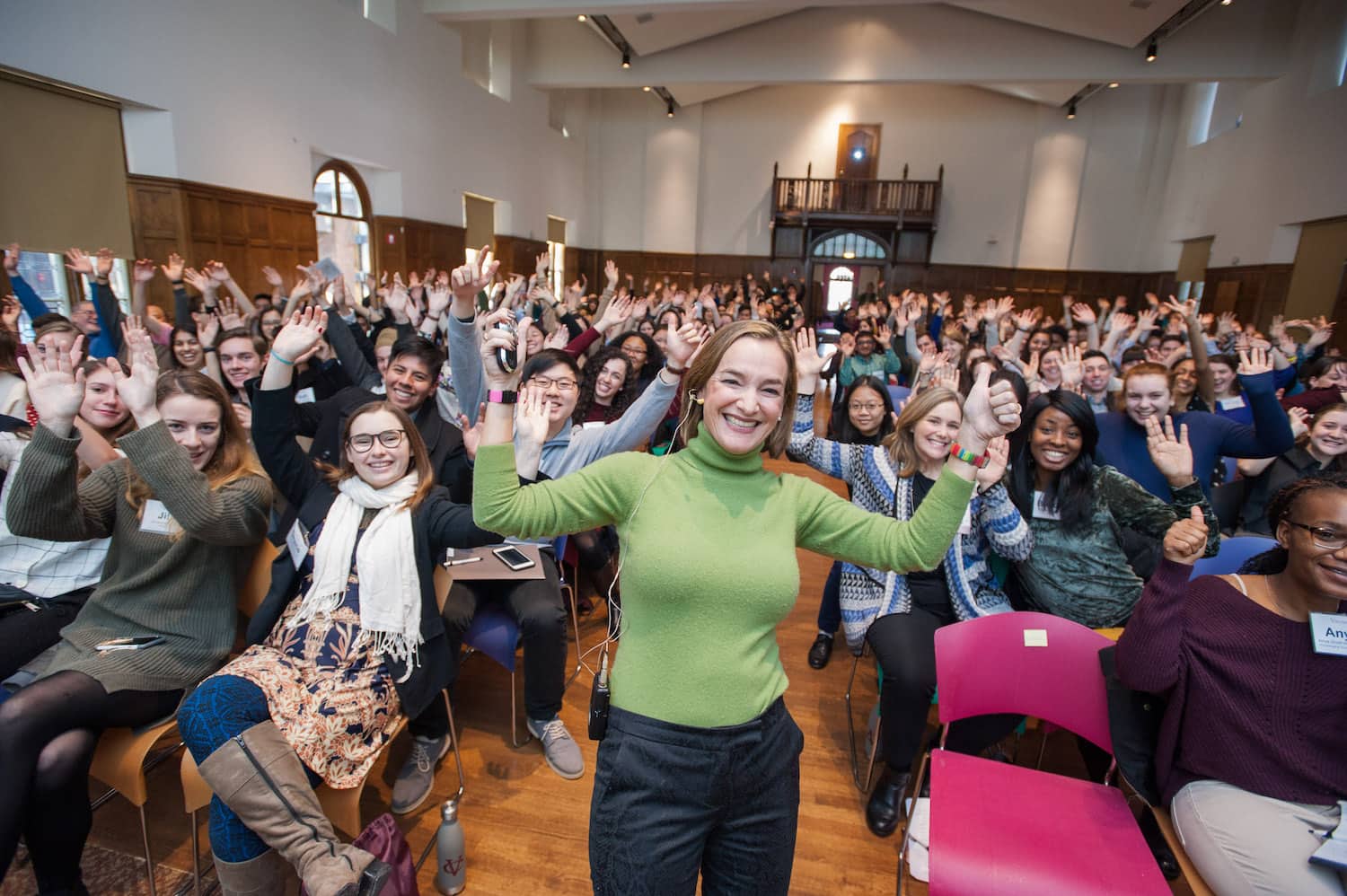 A person with long light brown hair wearing a green sweater and blue pants stands in front of a seated audience in a large room with their hands raised, smiling at the viewer.