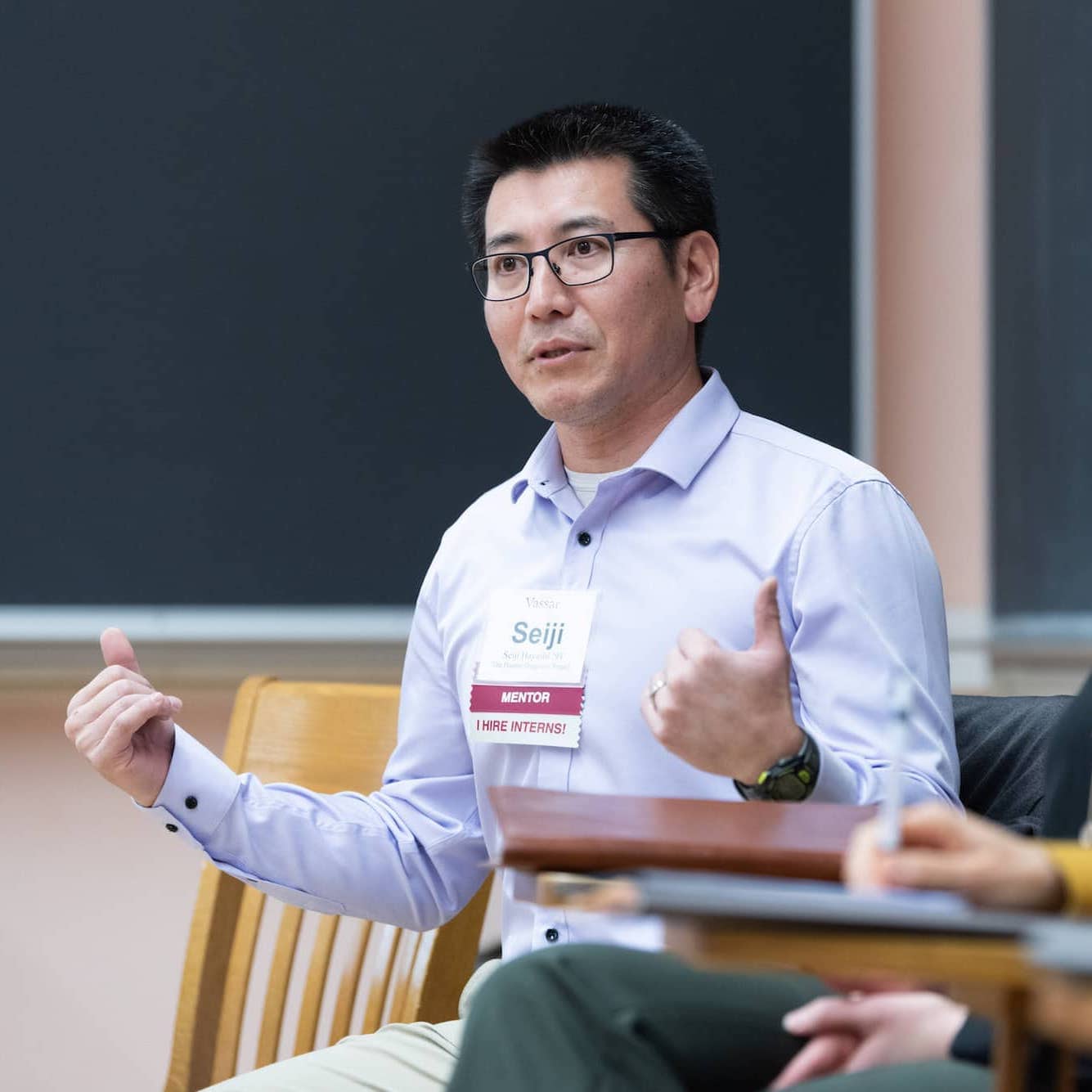 A person with short dark hair, glasses, and a purple button-down shirt, wearing a name tag that says Seiji, sits in a chair in a classroom talking.