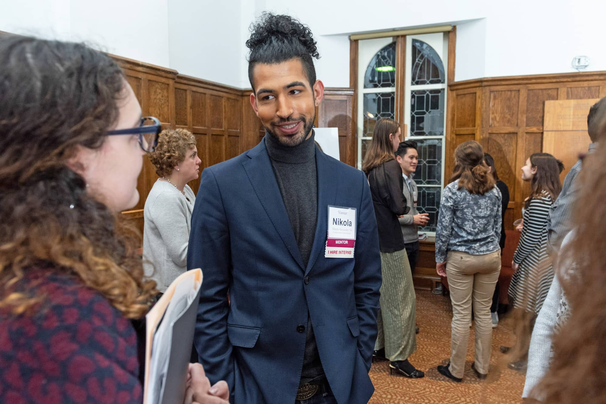 One person with their curly hair up wearing a blue blazer and grey turtle neck stands next to a person who has curly hair and glasses as they talk at an event.