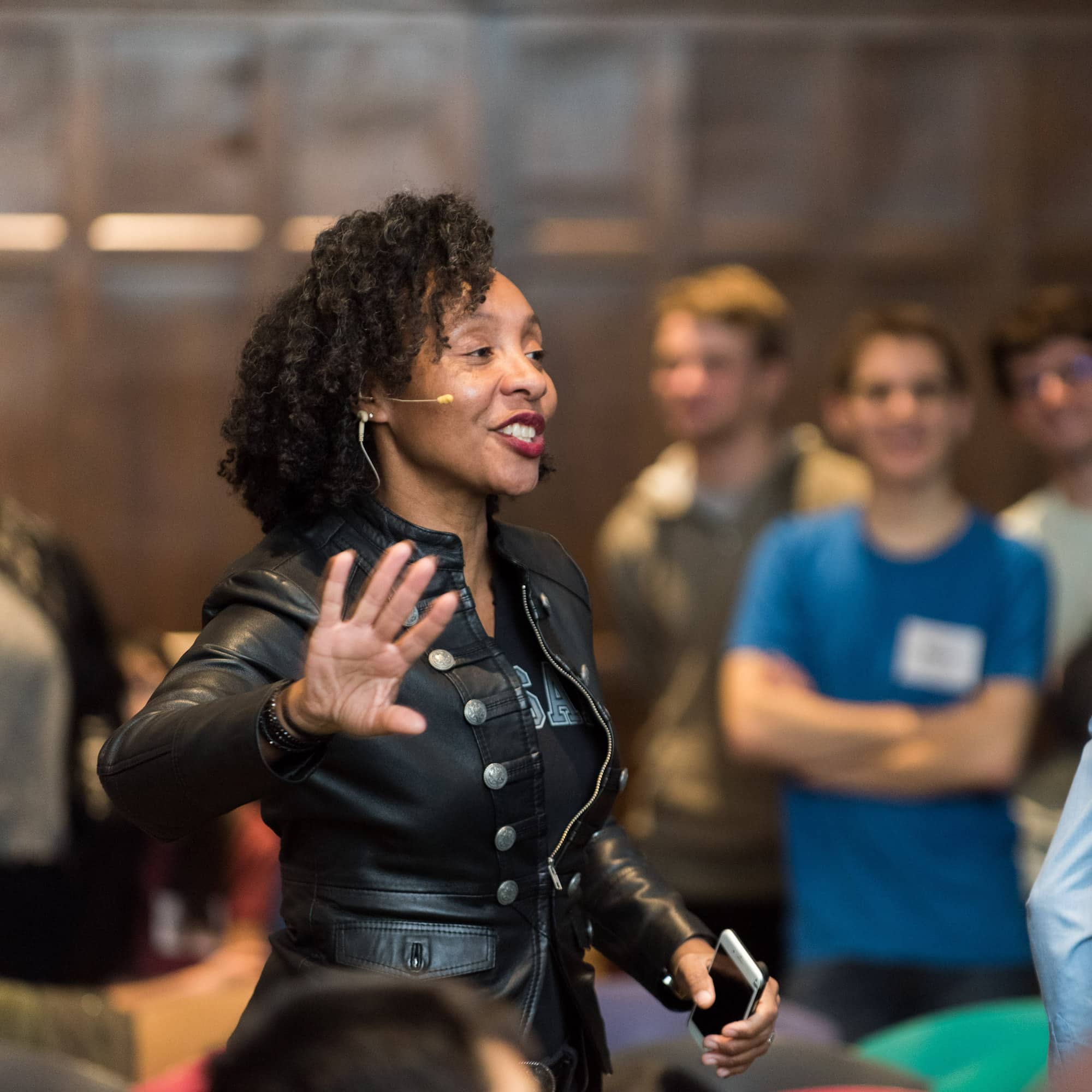 A person with short dark hair wearing a black jacket and a microphone, gesturing and speaking to a group.