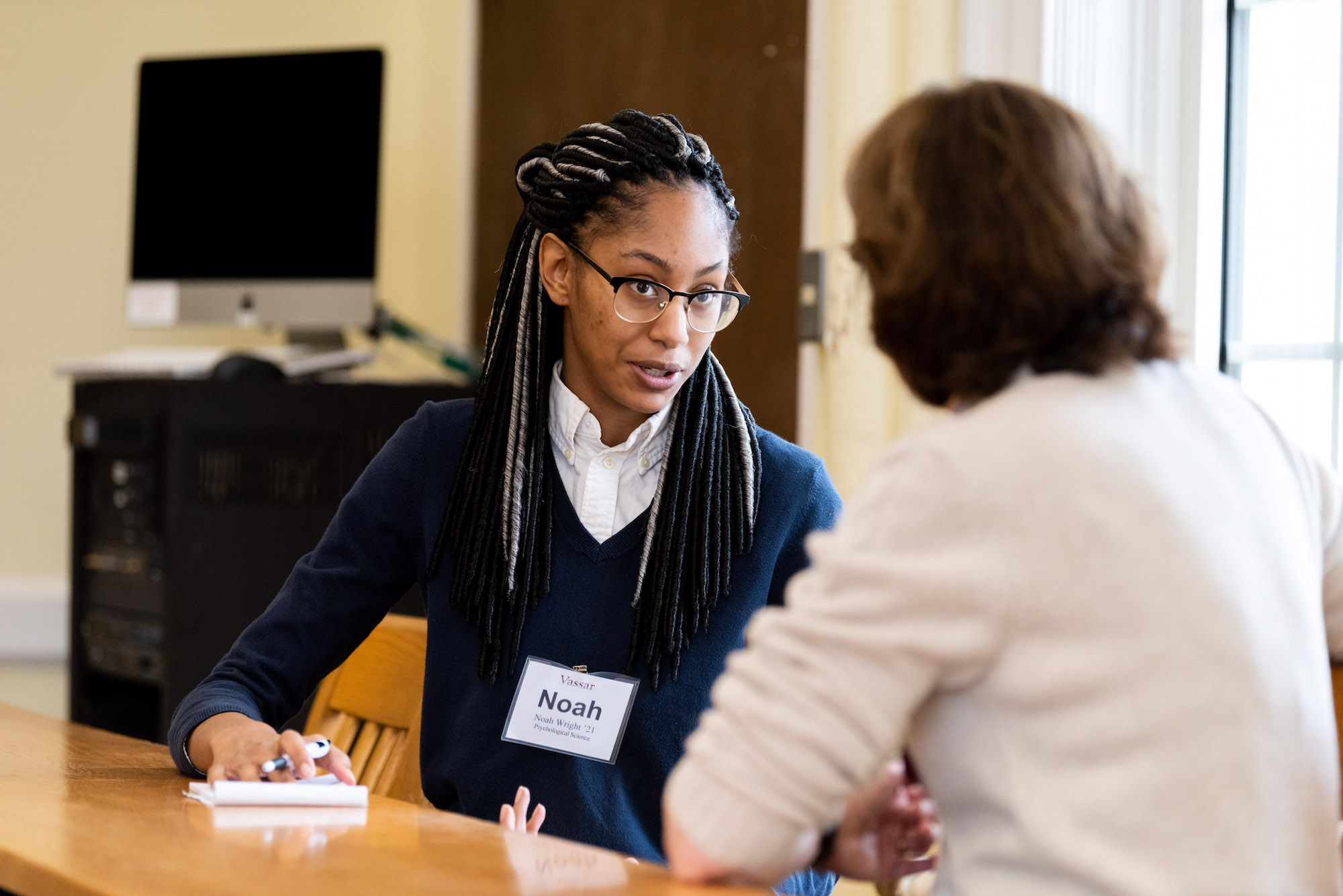 One person with long dark hair, glasses, and a blue sweater with a white collar sits at a table conversing with another person in a white sweater with their back to the viewer.