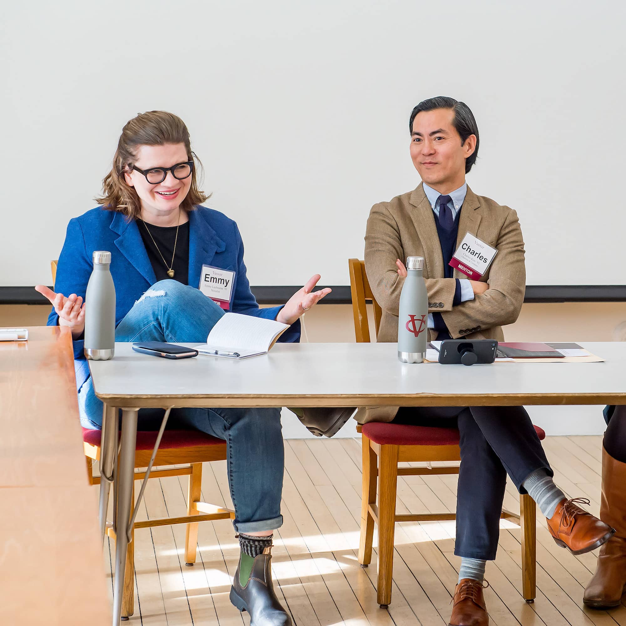 Two people sit at a table, one wearing a blue blazer and jeans, the other wearing a tie and beige suit jacket, both wearing name tags, while one of them speaks to someone outside of view.