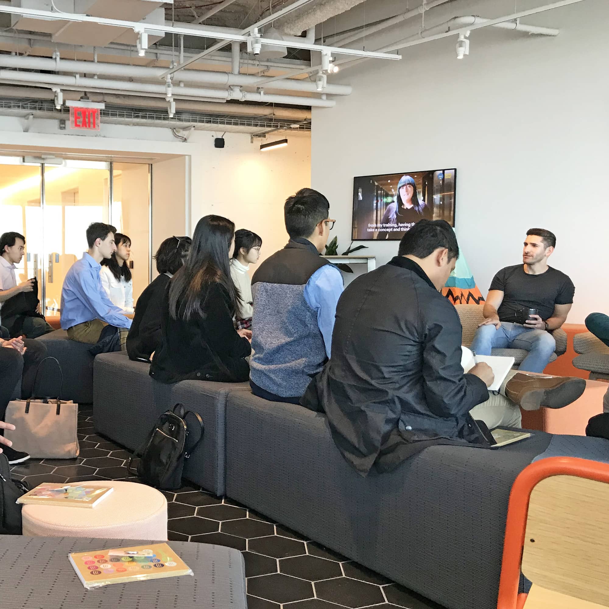 A group of students sit on cushioned chairs facing a video screen and a person in a black shirt and blue jeans seated in a chair.