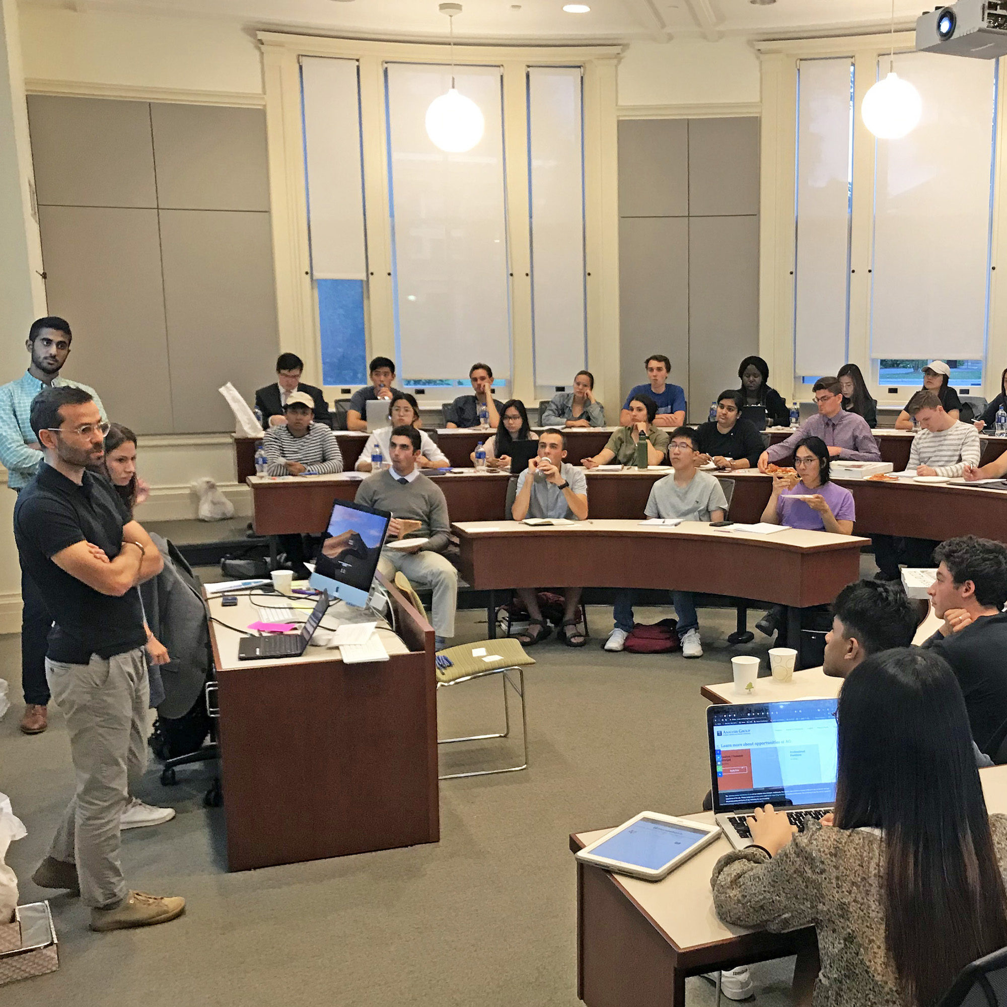 A group of people seated behind desks in a classroom facing a standing group of three people speaking.