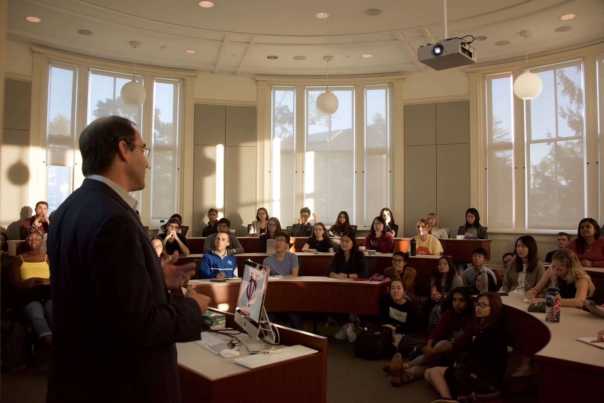 A person with short dark hair and a formal jacket speaks to a seated group of people in a classroom from behind a podium.