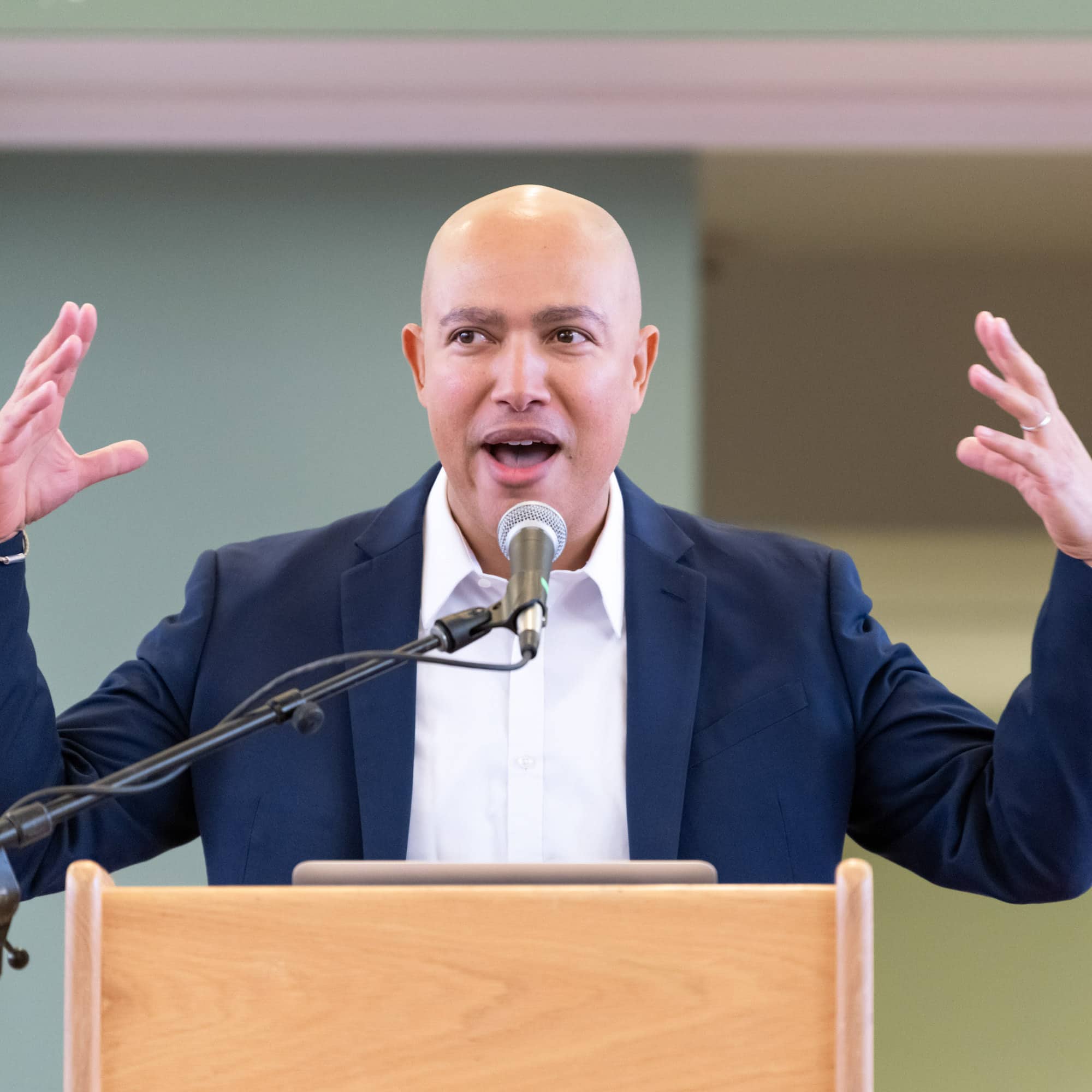 A person who is bald wearing a formal blue jacket and white collared shirt stands in front of a microphone speaking and gesticulating.