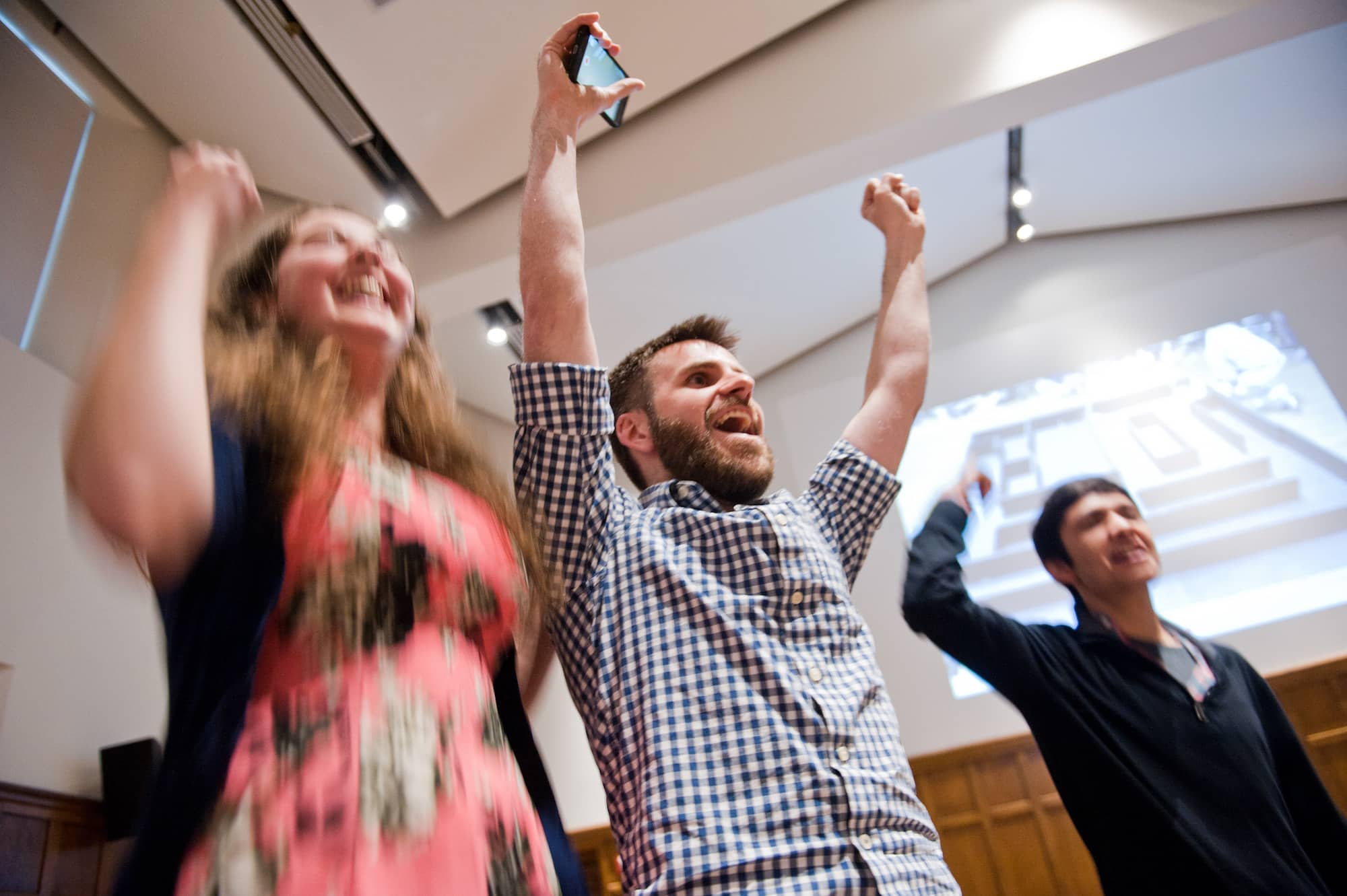 Three smiling people excitedly putting their arms in the air, standing in an indoor space.