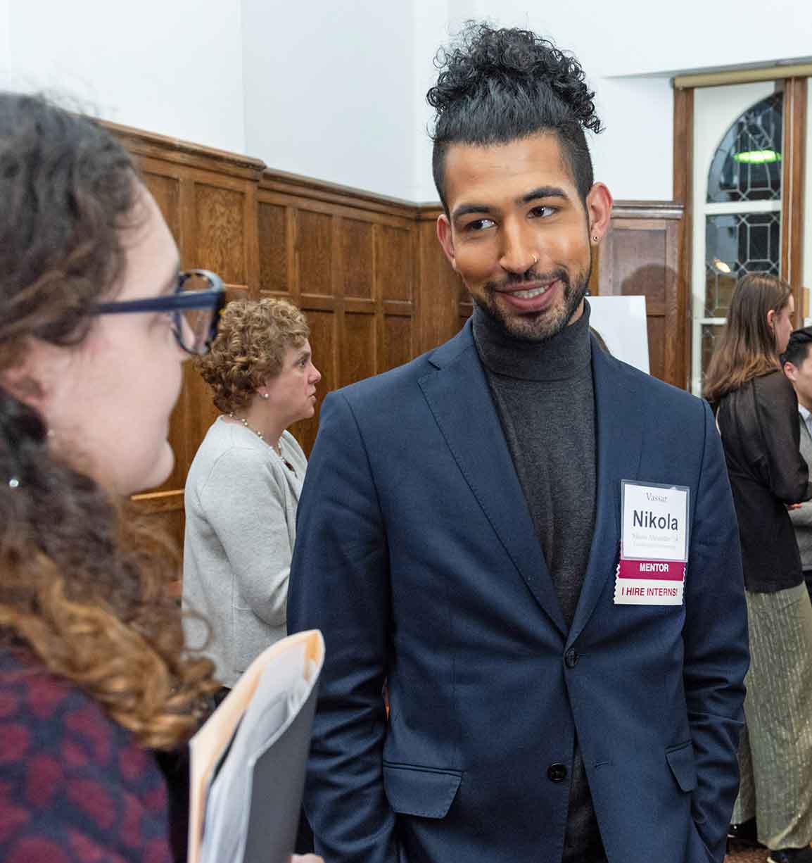 One person with their curly hair up wearing a blue blazer and grey turtle neck stands next to a person who has curly hair and glasses as they talk at an event.