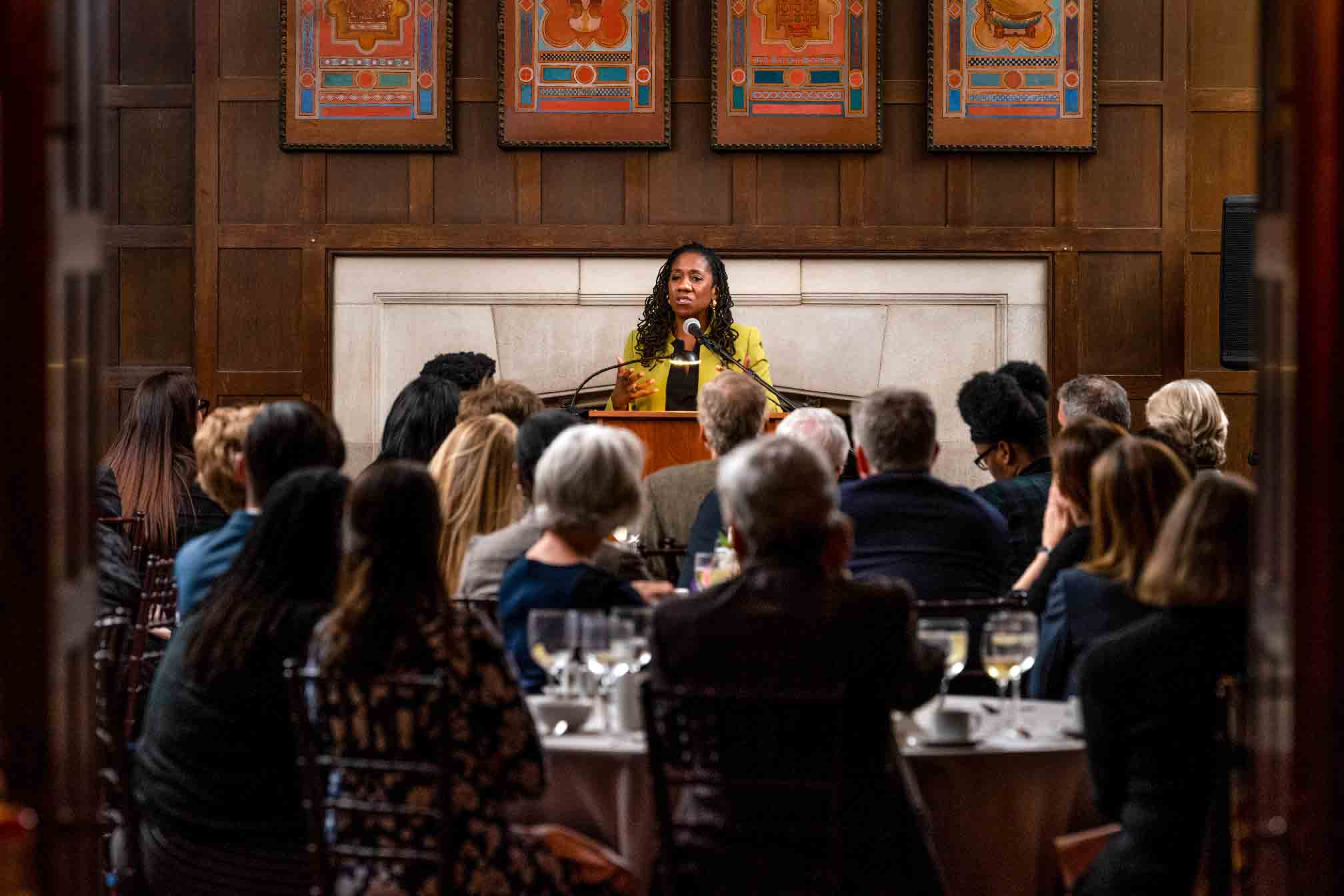 A person with long dark hair, yellow jacket, and a black shirt, Sherrilyn Ifill, stands behind a podium, speaking to a seated audience at a Vassar event.