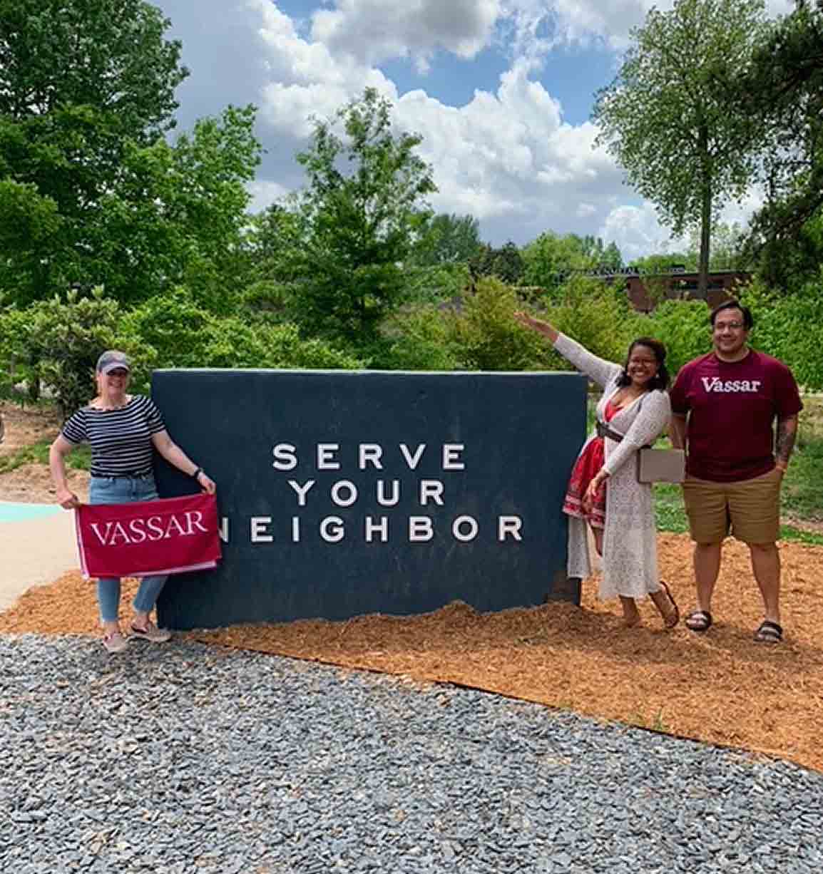 People wearing and holding Vassar merchandise standing beside a sign that reads, Serve Your Neighbor.