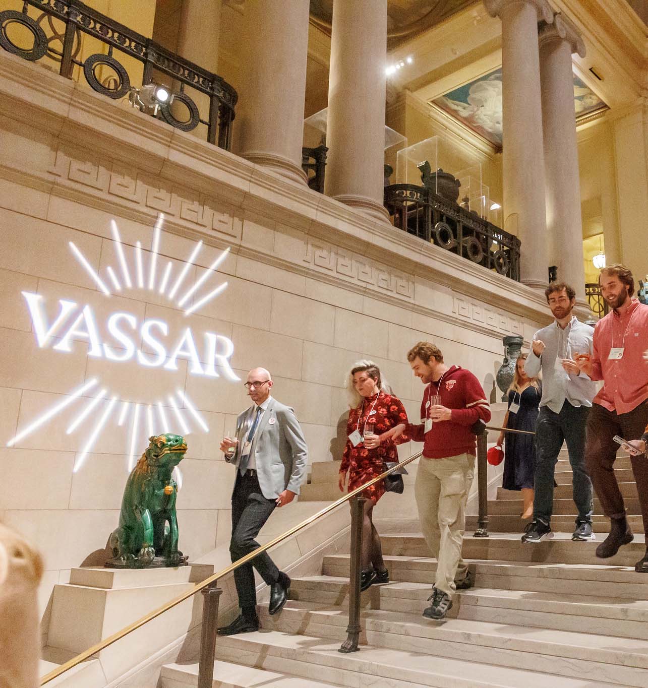 People walking down a large staircase in a museum with a projected image of a Vassar Fearlessly Consequential campaign logo project on a wall.