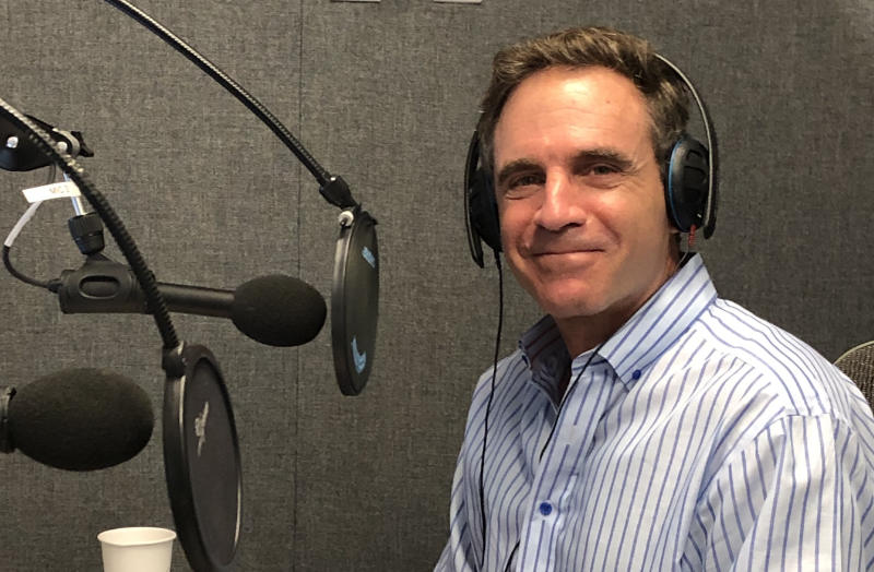 A person sits in a soundproof studio wearing over-ear headphones and a blue-striped shirt. They are surrounded by three microphones with foam covers and pop filters on adjustable arms. The gray acoustic panels on the wall and a white paper cup in the lower-left corner suggest a controlled recording environment. Lighting is even and soft, with minimal shadow.