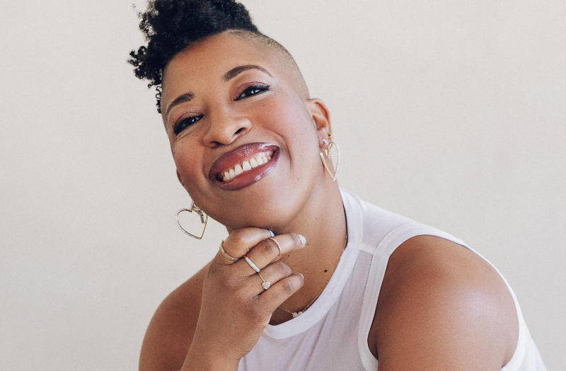 A person poses in front of a plain off-white background with one hand resting under their chin. They wear a sleeveless white shirt, heart-shaped hoop earrings, and several rings. Their hair is styled in a tall, natural curl with one side closely shaved. Bright, even lighting enhances facial highlights and gives the image a clean, high-contrast look.