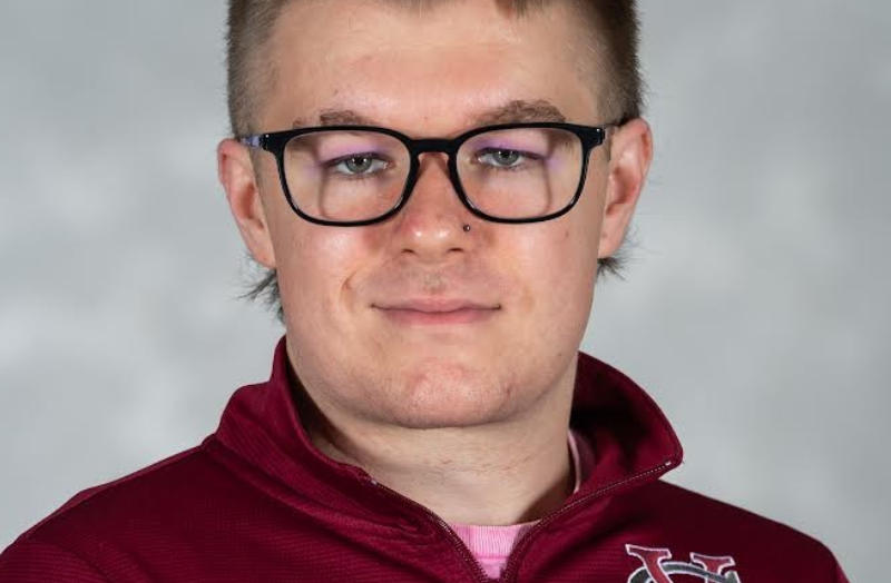 A person with short, brushed-back hair and rectangular glasses looks directly at the camera. They wear a maroon athletic pullover with a white logo on the chest and a light pink shirt underneath. The background is a soft gradient of gray tones with gentle focus falloff. The lighting is uniform, evenly illuminating the face and clothing.