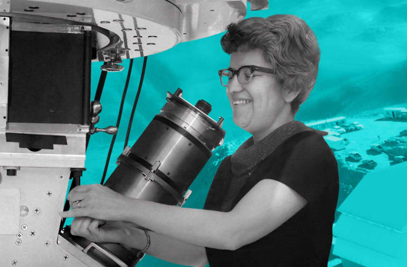 A person operates a large cylindrical mechanical device mounted under a complex metal structure. They wear glasses and a short-sleeved top, with short, curled hair and a bracelet on their wrist. The background is digitally edited in cyan and features a faint landscape with buildings and mountainous terrain. The foreground remains in grayscale, emphasizing the machinery and figure.