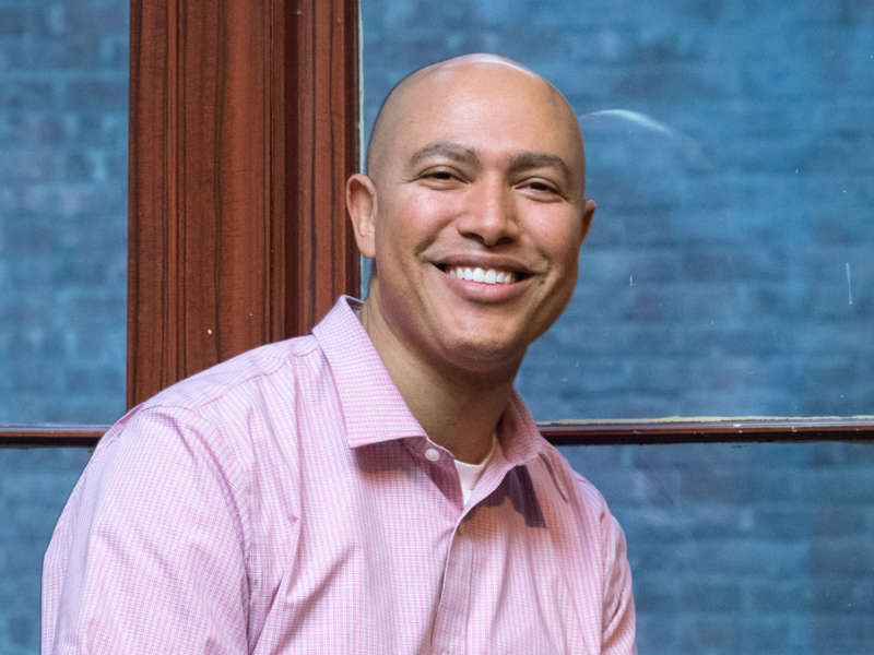 A smiling figure sits beside a large window, framed by rich wooden panels that echo a warm tone. The figure wears a light pink, checkered shirt and appears relaxed, leaning slightly forward. Outside the window, a textured blue backdrop suggests a building or urban environment. Soft, diffused light illuminates the scene, enhancing the inviting atmosphere.