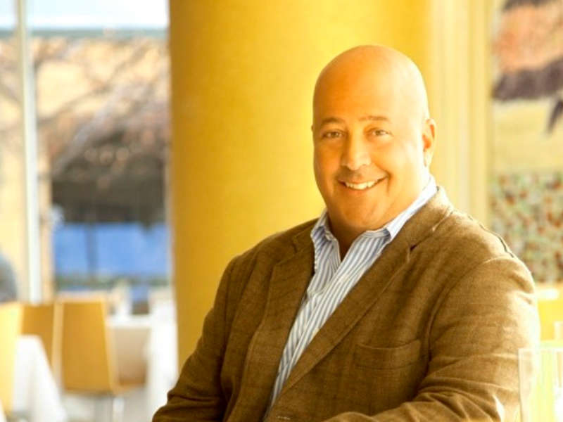 A smiling figure is seated indoors, positioned to the left of the frame. The background features a warm yellow wall and glimpses of a softly blurred outdoor setting, creating a calm atmosphere. The soft lighting highlights the figure’s expression and attire, consisting of a light-colored shirt beneath a textured brown blazer. Minimal furnishings, such as light-colored chairs and tables, are visible in the background, enhancing the inviting environment.