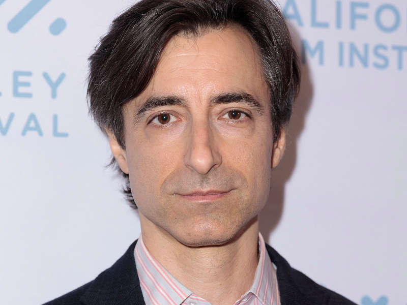 A close-up portrait features an individual with dark, tousled hair and a neutral expression. The subject is wearing a dark blazer over a light-striped shirt, which introduces subtle contrast against the light background. Behind, text related to a film festival is partially visible, rendered in soft blue hues. The lighting is even and highlights the subject’s features against the plain backdrop, creating a professional and polished appearance.