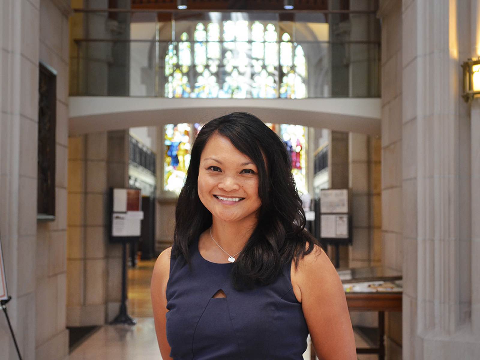Michele Tugade ’95, a person with long black hair and a blue dress, stands in the Vassar Library, a stone building with a stained glass window.