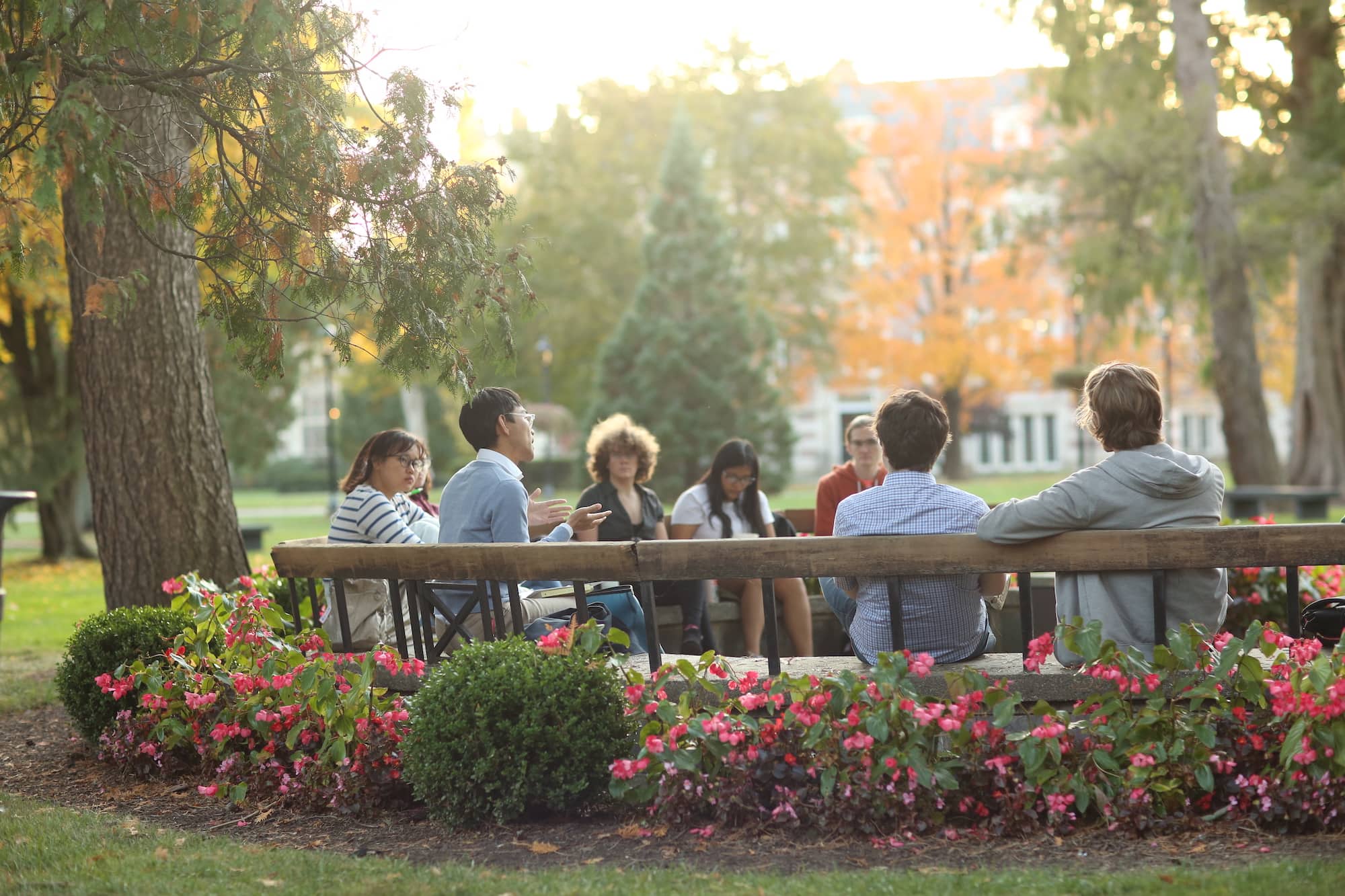 A group of people sitting outside on a large circular wood bench with flowering shrubs planted behind the bench.