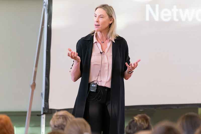 A person with mid-length blonde hair in a black sweater and pink button-down shirt stands in front of a seated audience speaking and gesturing.