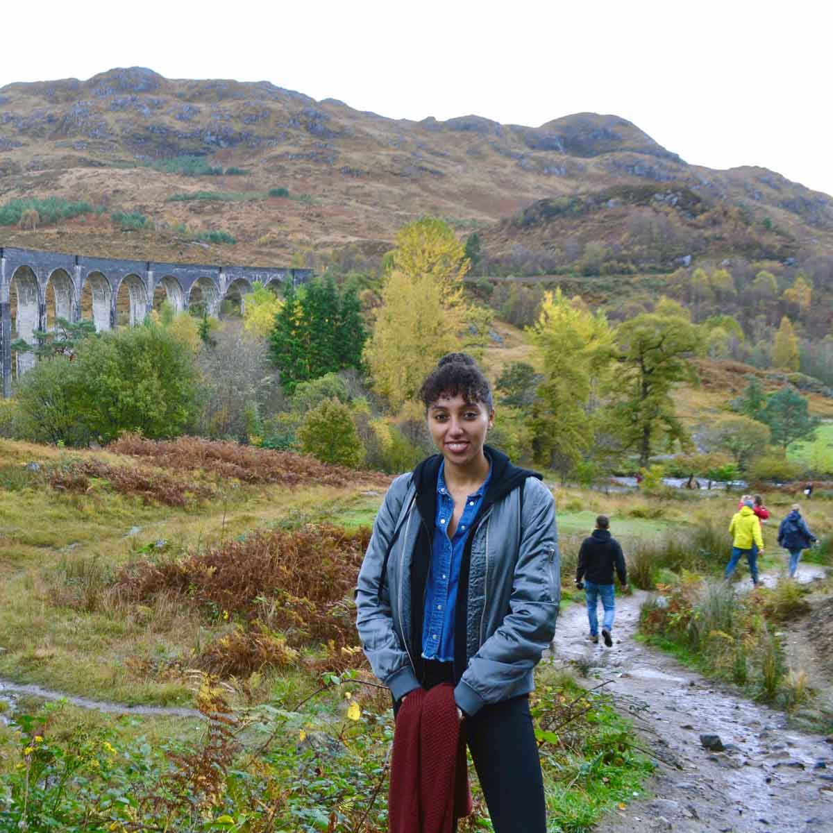 A person with dark hair wearing a blue jacket and dark pants is standing in front of an aqueduct surrounded by greenery.