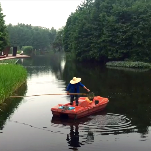 A person wearing a straw hat stands holding a net on a pole in a small orange boat on a river.