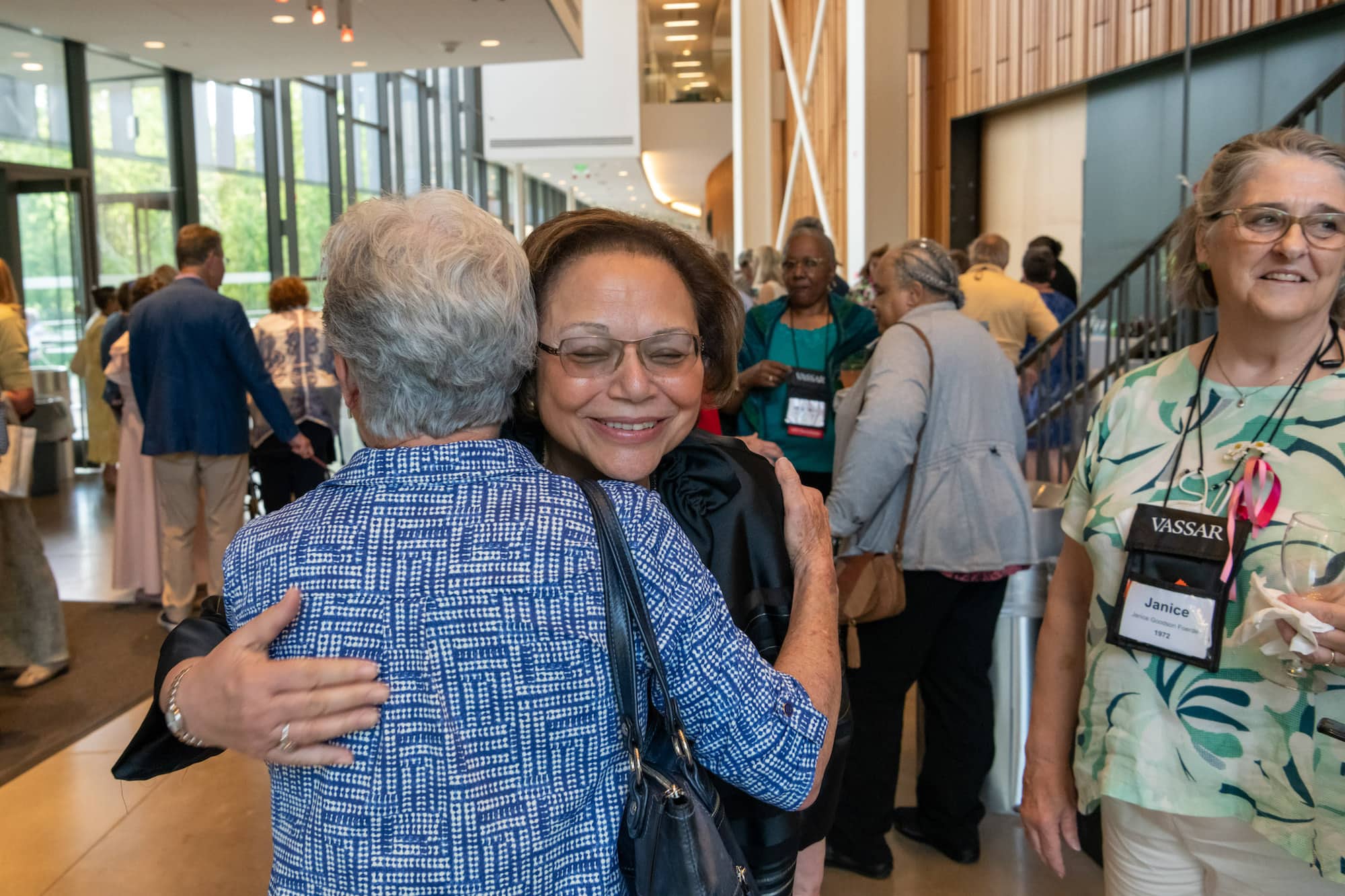 Two people, one with short white hair wearing a blue shirt and the other with short dark hair, wearing a dark shirt embrace each other at a crowded event.