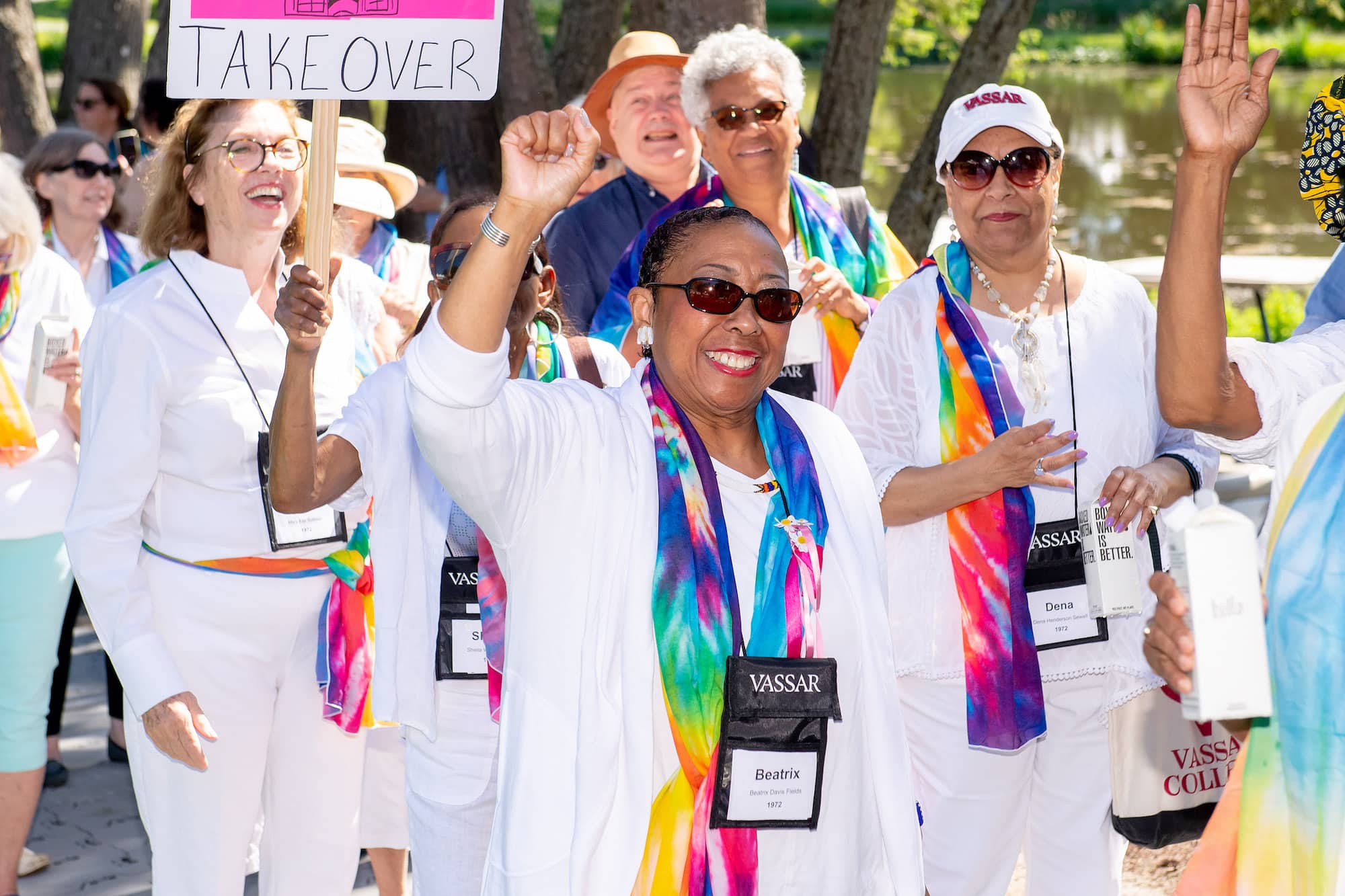 A group of people in a procession wearing white clothing and multicolored scarves raise their hands in the air and smile at the viewer.