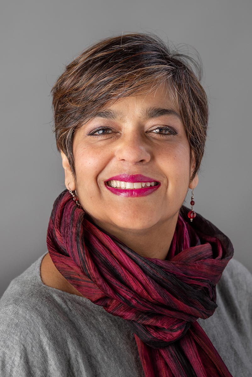 A person with short brown hair and pink lipstick wearing a gray shirt and black and red scarf against a gray background, smiling at the viewer.