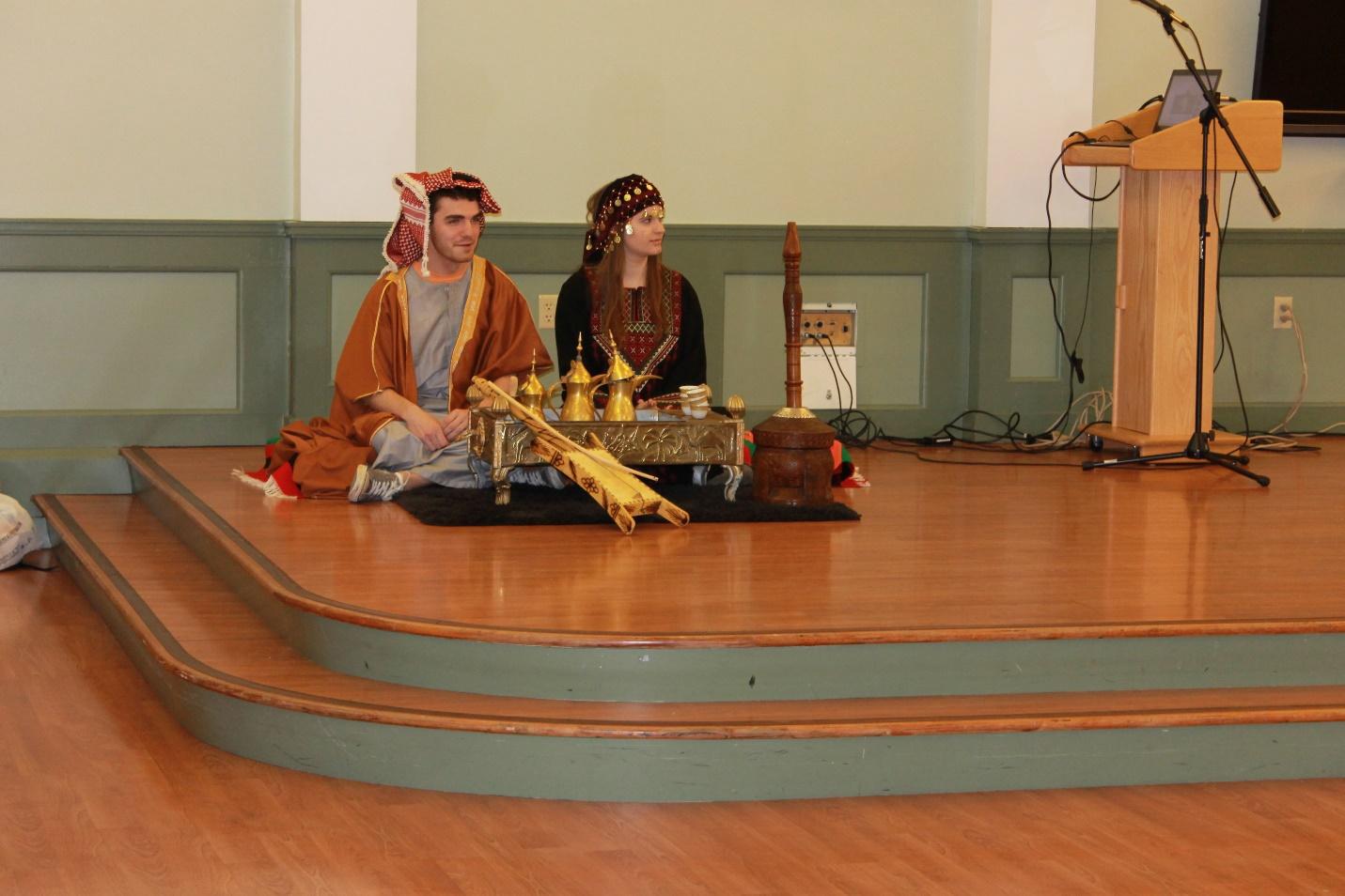 Two students in traditional Arabic clothing sit on a hardwood stage
