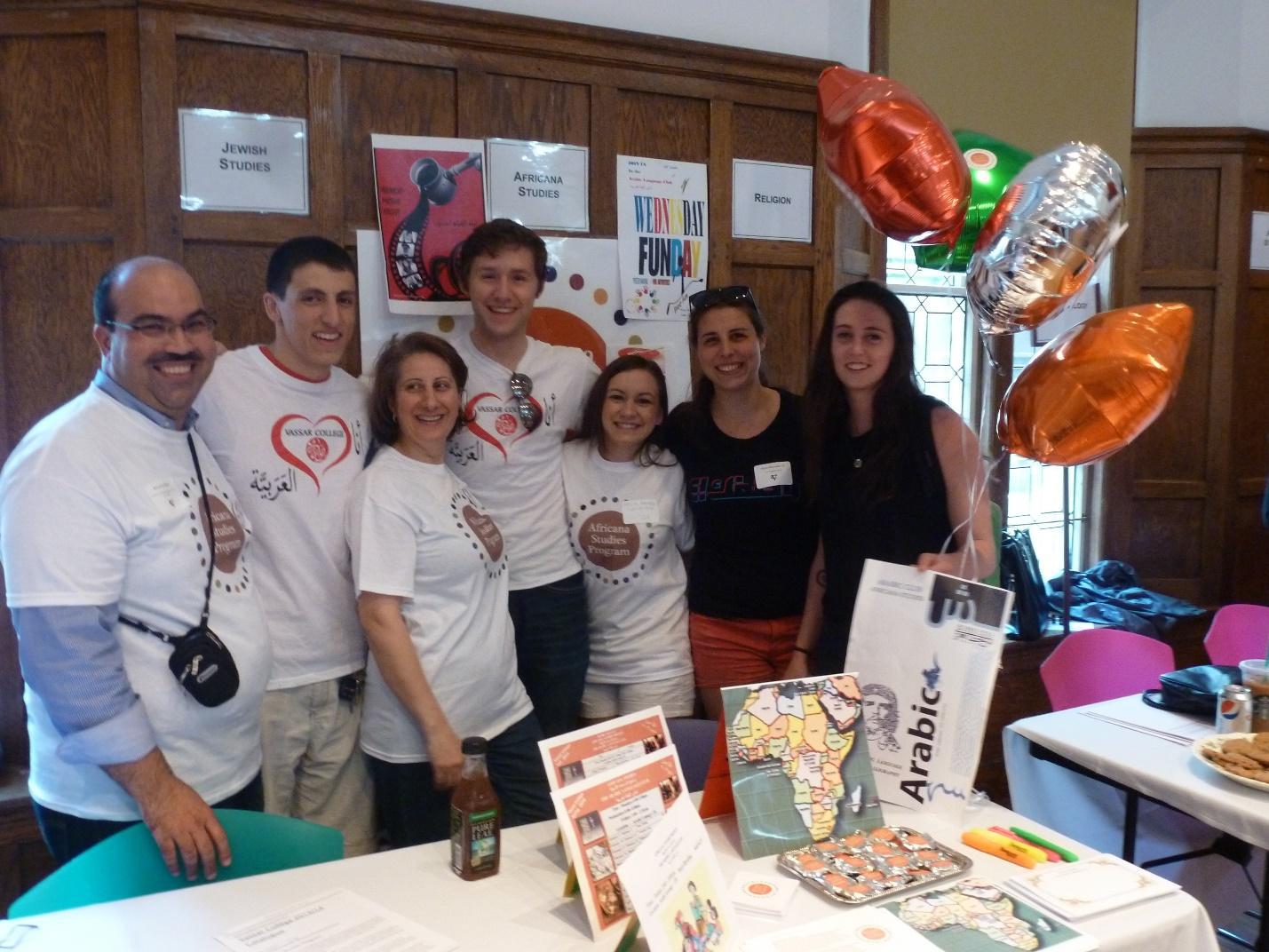 A group of students and faculty stand behind a table with department literature on it, smiling at the camera