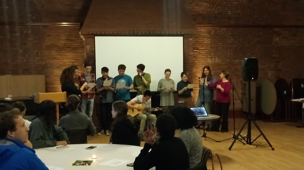 A group of students stand at the front of a large room, holding sheets of paper and singing