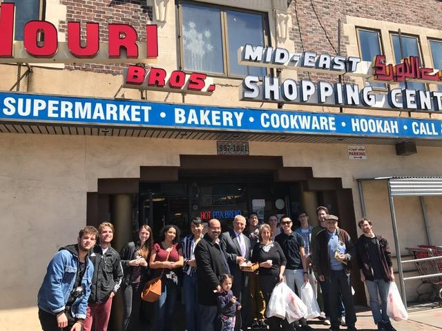 A large group of students and faculty stand in front of a supermarket selling Middle Eastern goods