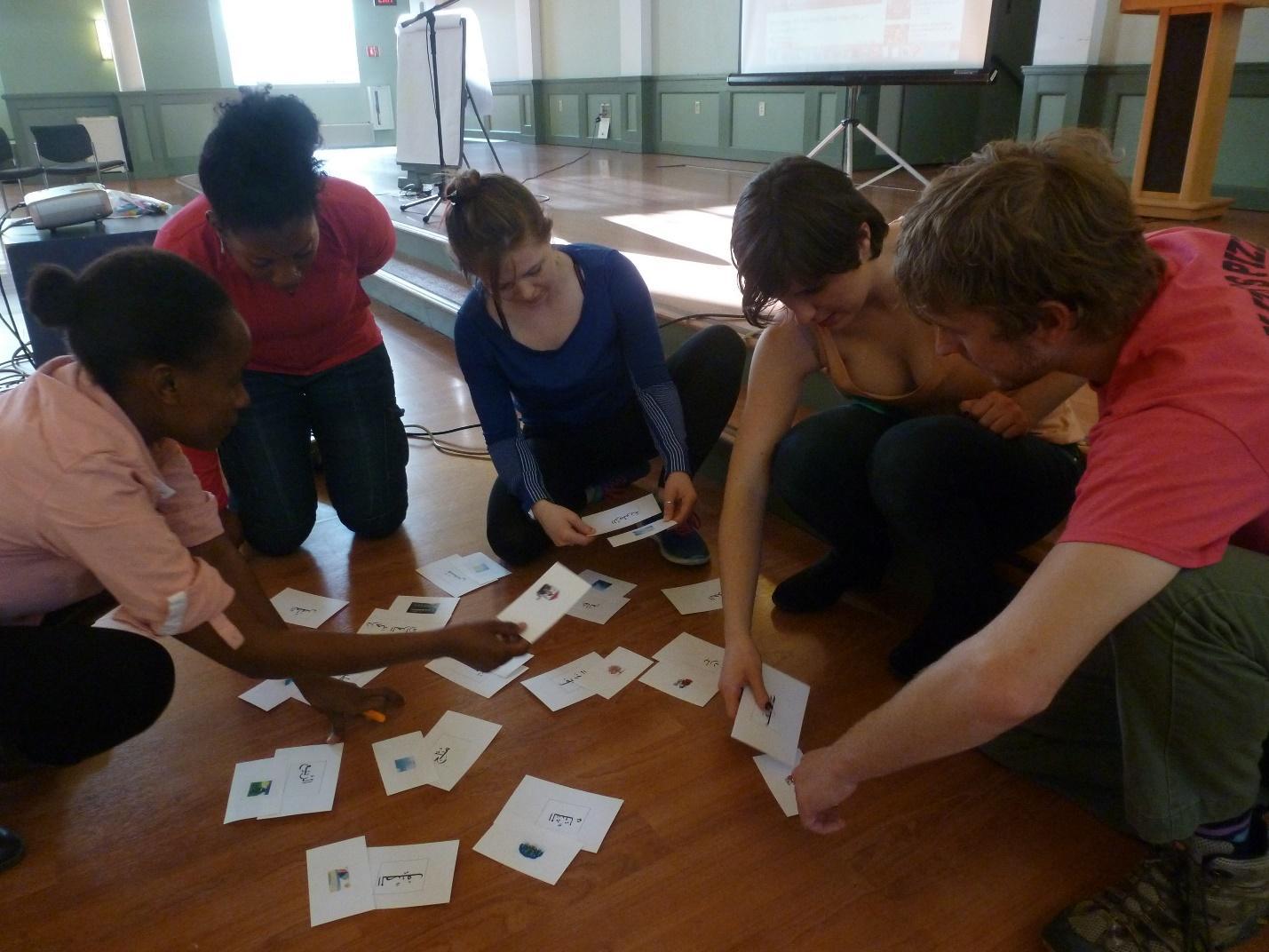 Several students sit on a hardwood floor, handling small paper cards.