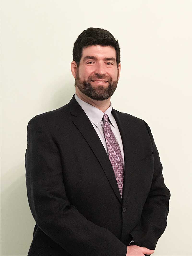 A person with thick dark brown hair, a beard, and a dark suit and lavender-patterned tie smiles at the viewer.