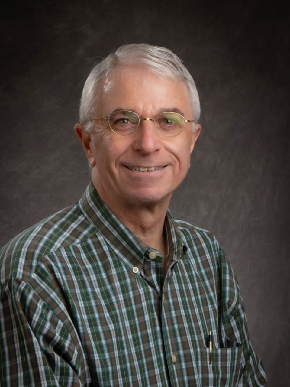A person with short gray hair, glasses, and a green checkered shirt smiles at the viewer.