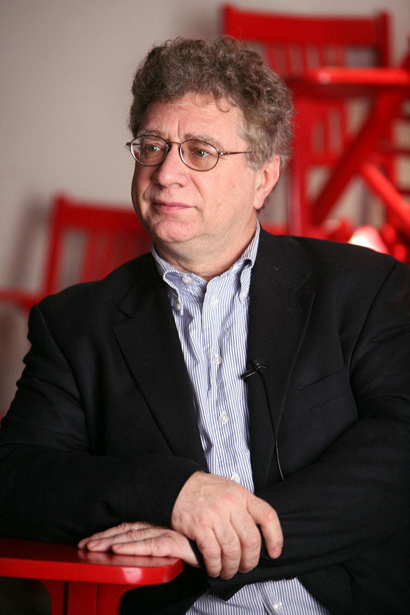 David A. Kennett sitting in a red chair wearing a white and blue striped shirt and black jacket. 