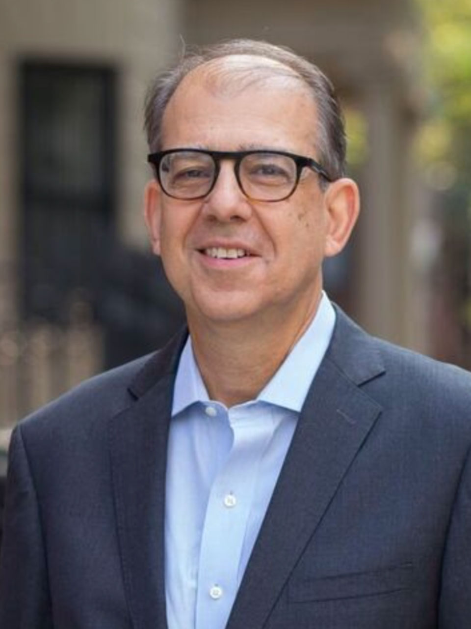 A person with thinning gray hair, glasses, and a gray formal jacket smiles at the viewer.