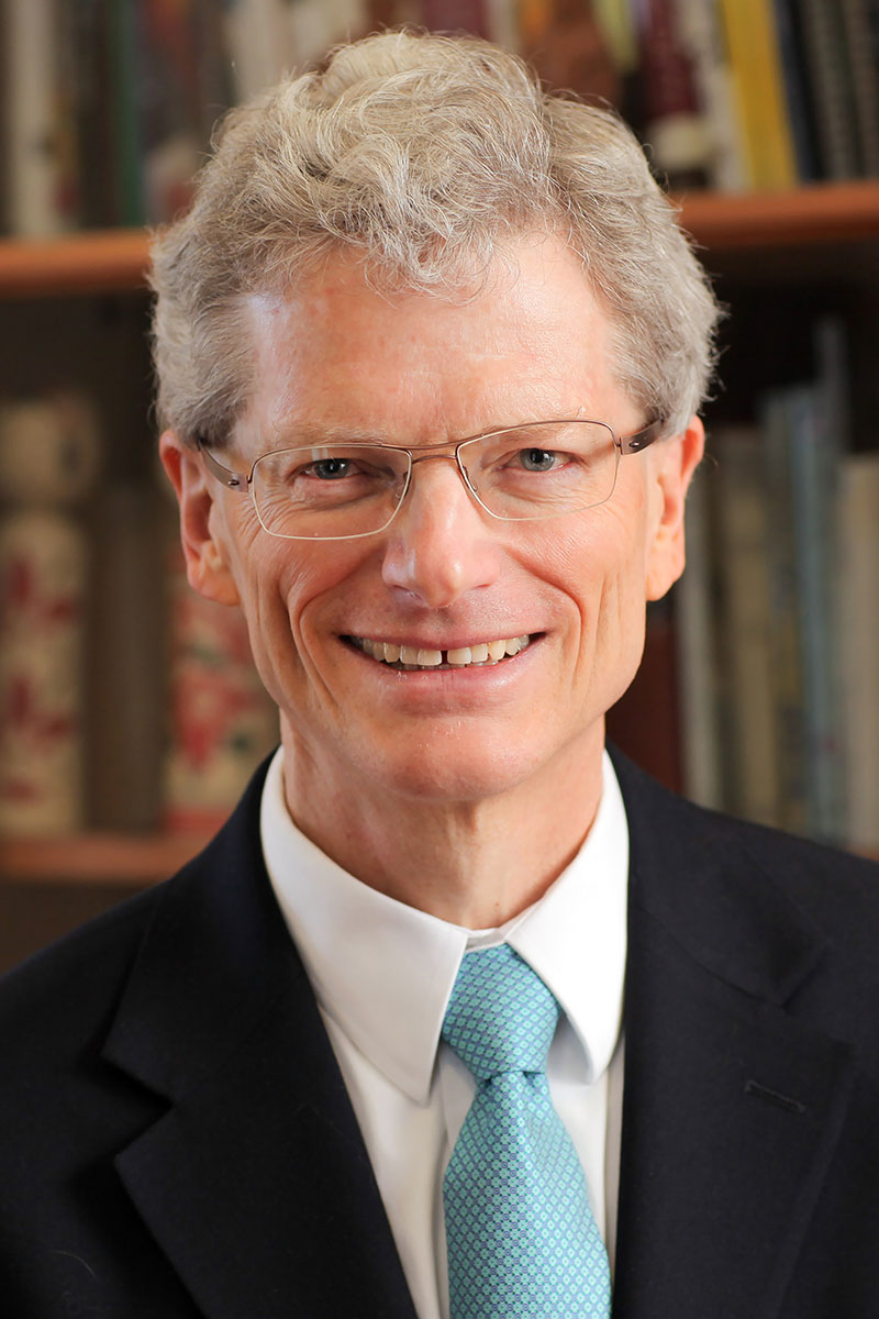 Jon Chenette wearing a white shirt, black jacket and light blue tie against a bookshelf background.