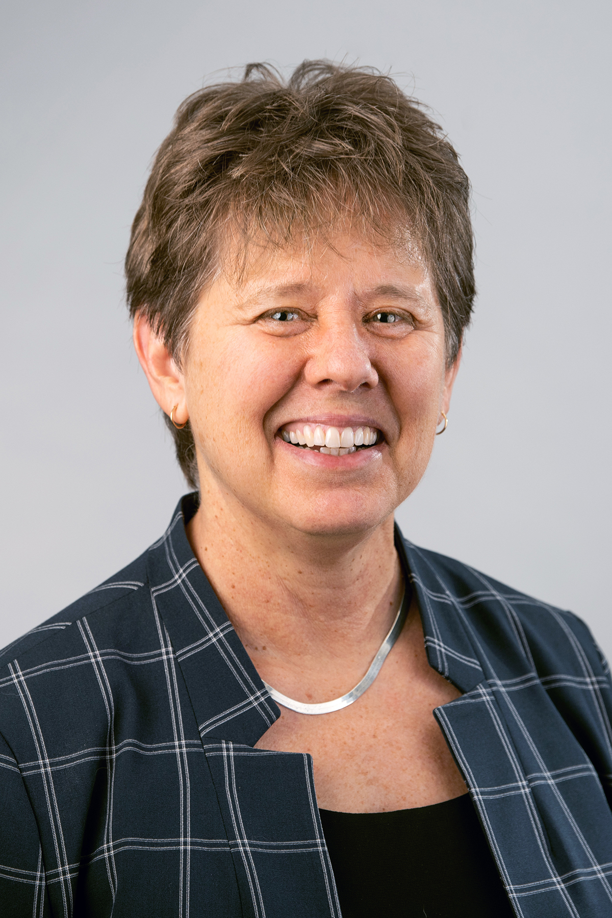 Jill S. Schneiderman wearing a black shirt, patterned dark blue shirt, and thick silver necklace against a light background.