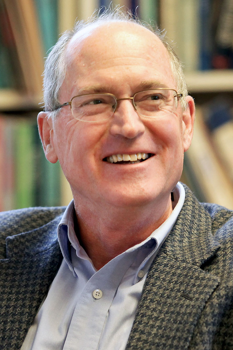 Stephen R. Rock wearing a light blue shirt and houndstooth jacket with a bookcase in the background. 