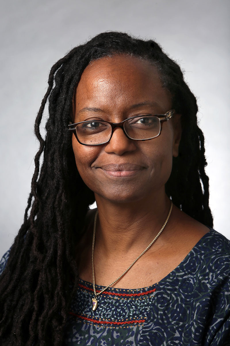 Candice Lowe Swift wearing a blue patterned shirt and foot charm necklace against a light gray background.