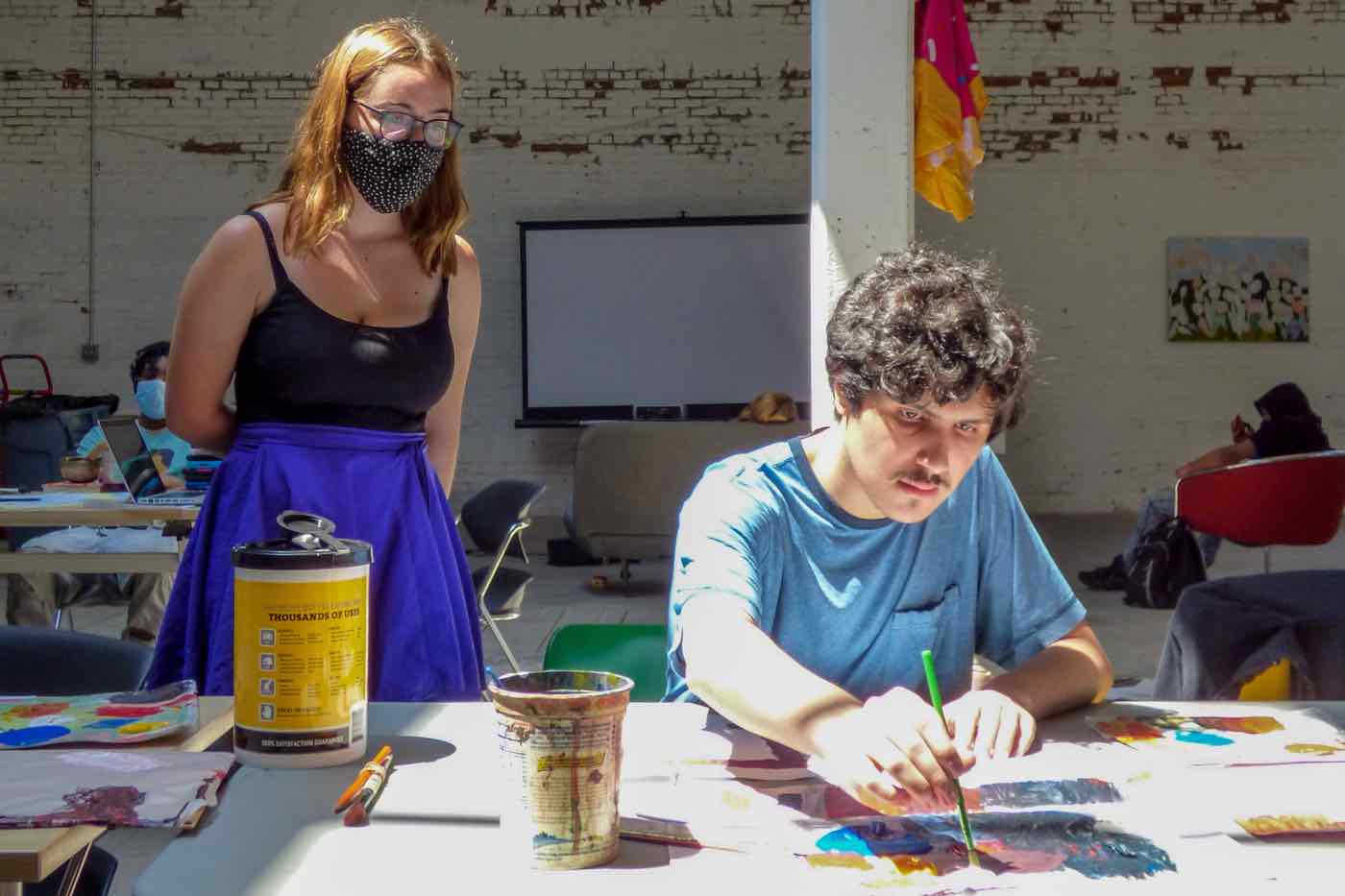 A female student looks at a male student painting a picture at a table.