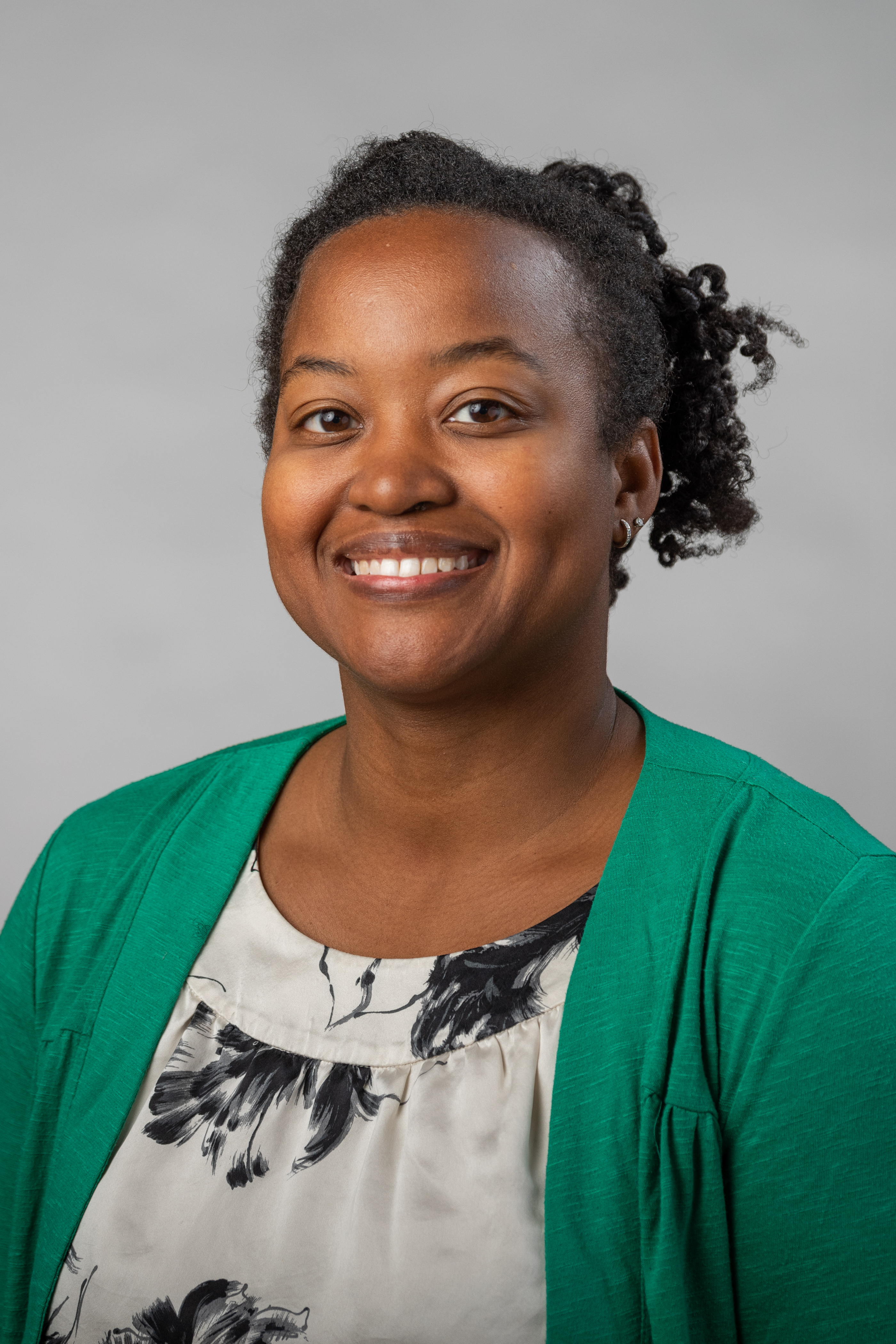 Kelli A. Duncan wearing a black and white patterned shirt and a green jacket with a gray background.