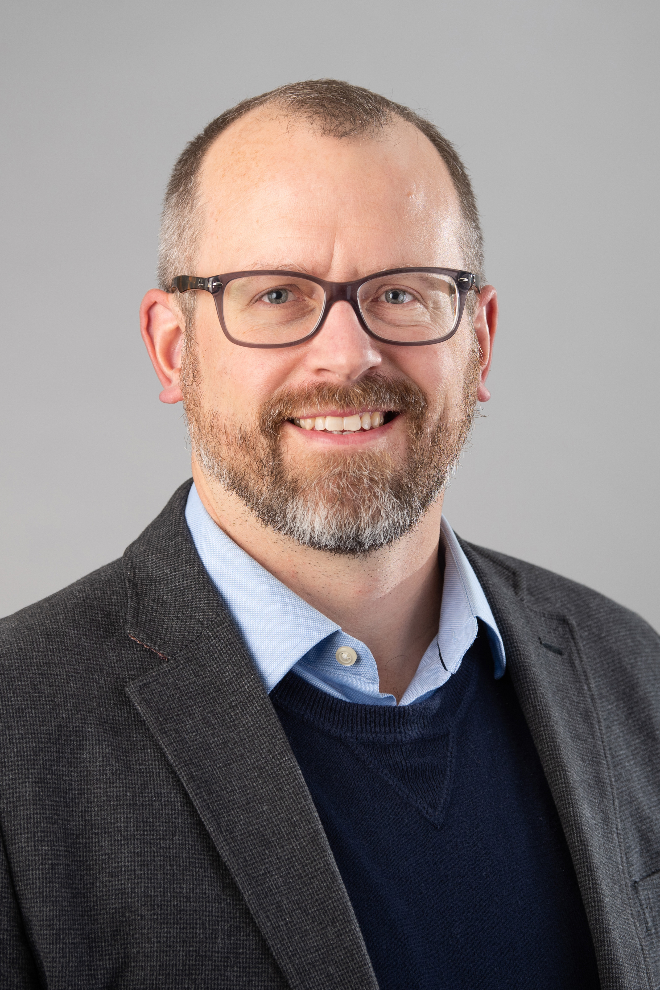 Zachary J. Donhauser wearing a light blue shirt, navy blue sweater and dark gray jacket against a gray background.