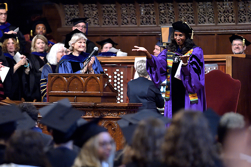 A room full of people in ornate formal gowns. A person with a purple gown speaks at a wood-paneled podium.