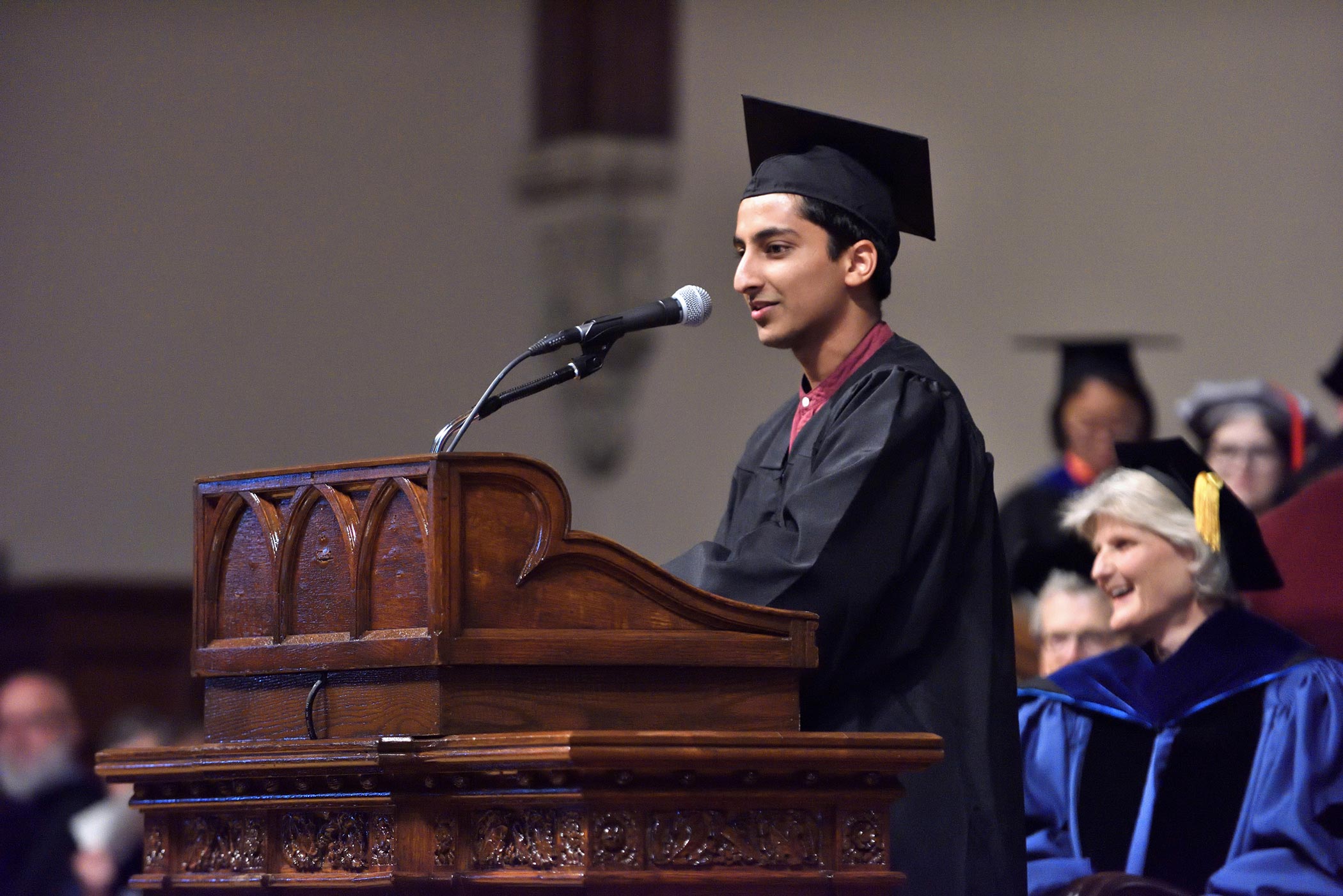 Anish Kanoria ’18 speaks at an ornate wooden podium. Kanoria wears a black robe and black cap.