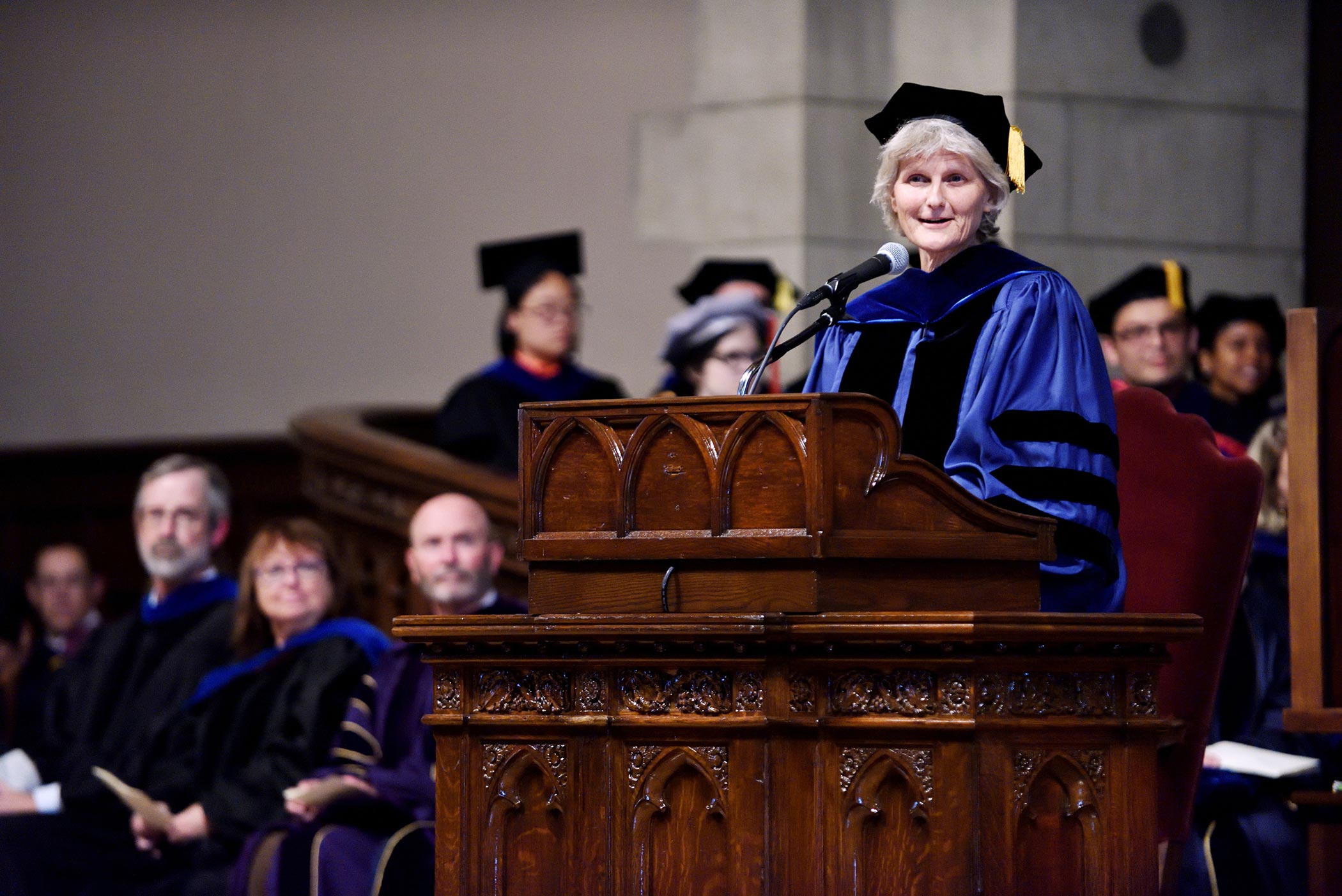 President Bradley speaks at an ornate wooden podium. Bradley wears a blue robe and black cap with gold tassel.