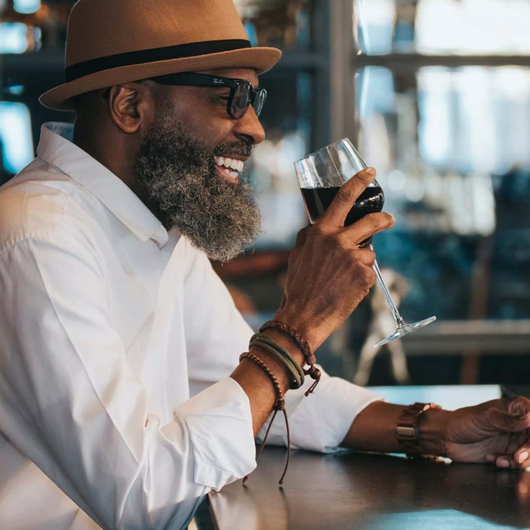 A person with a black beard, a brown hat, sunglasses, and a white shirt with a collar drinks a glass of wine at a bar.